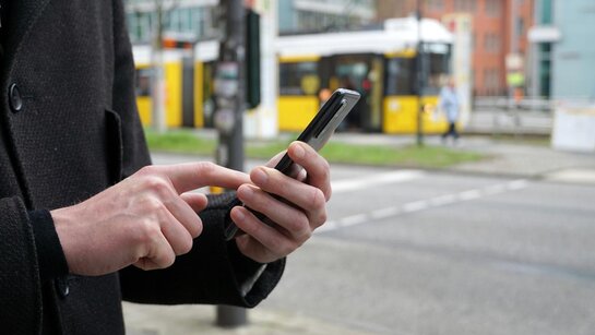 Smartphone mit Hand im Vordergrund, gelbe Tram im Hintergrund